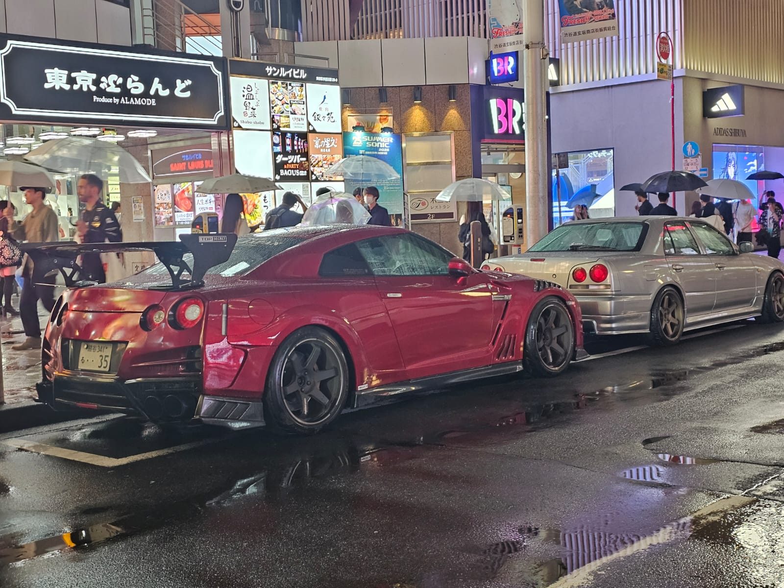 Red Nissan GT-R and silver Nissan Skyline R34 parked together on a rainy Shibuya street at night