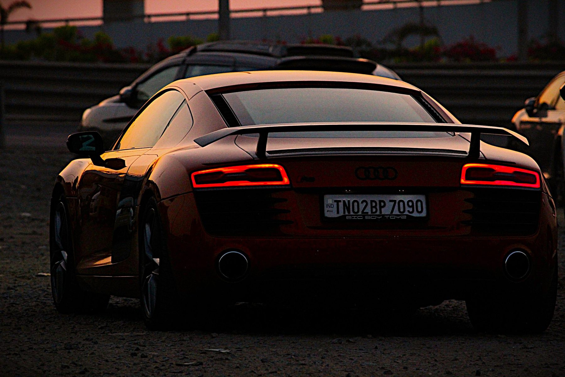 Bronze Audi R8 rear three-quarter view at sunset with taillights lit, next to a dealership sticker reading Big Boy Toyz