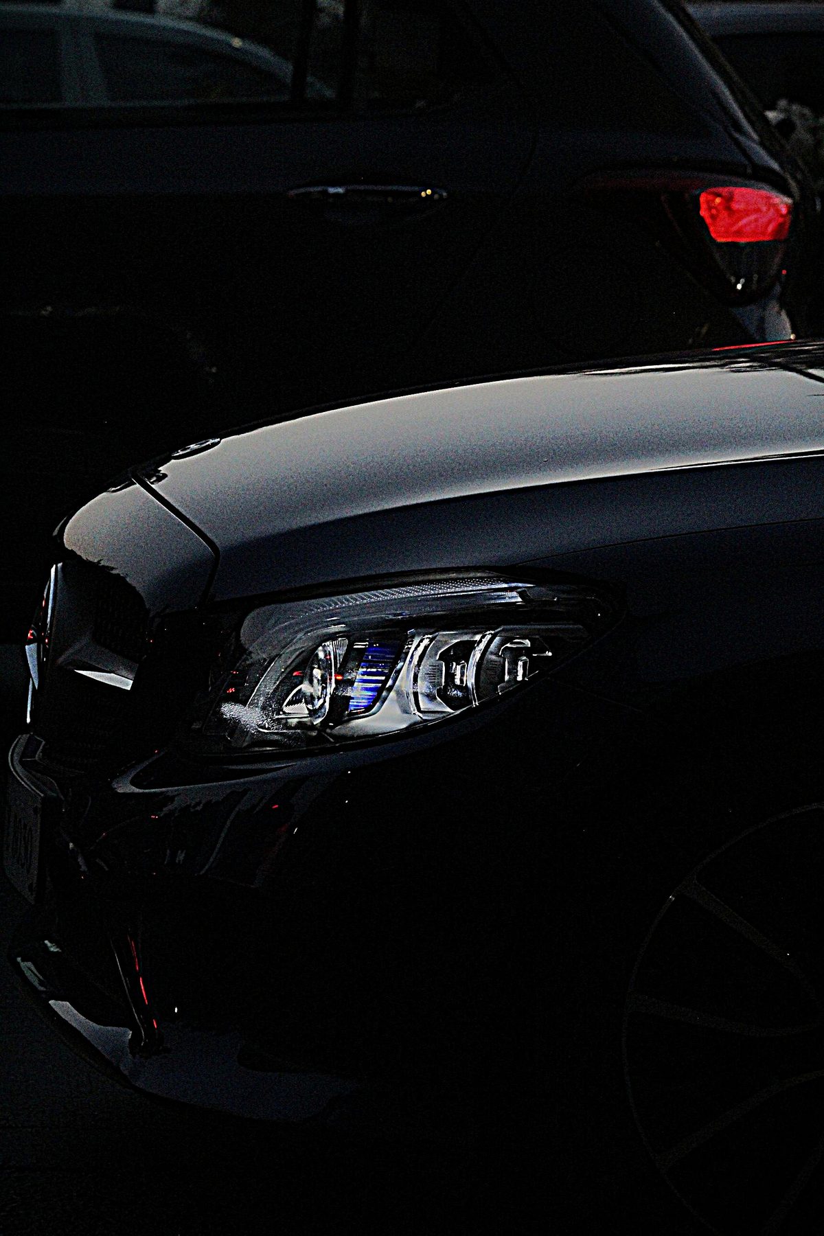 Close-up of a black Mercedes-Benz headlight at night with a red taillight glowing in the background
